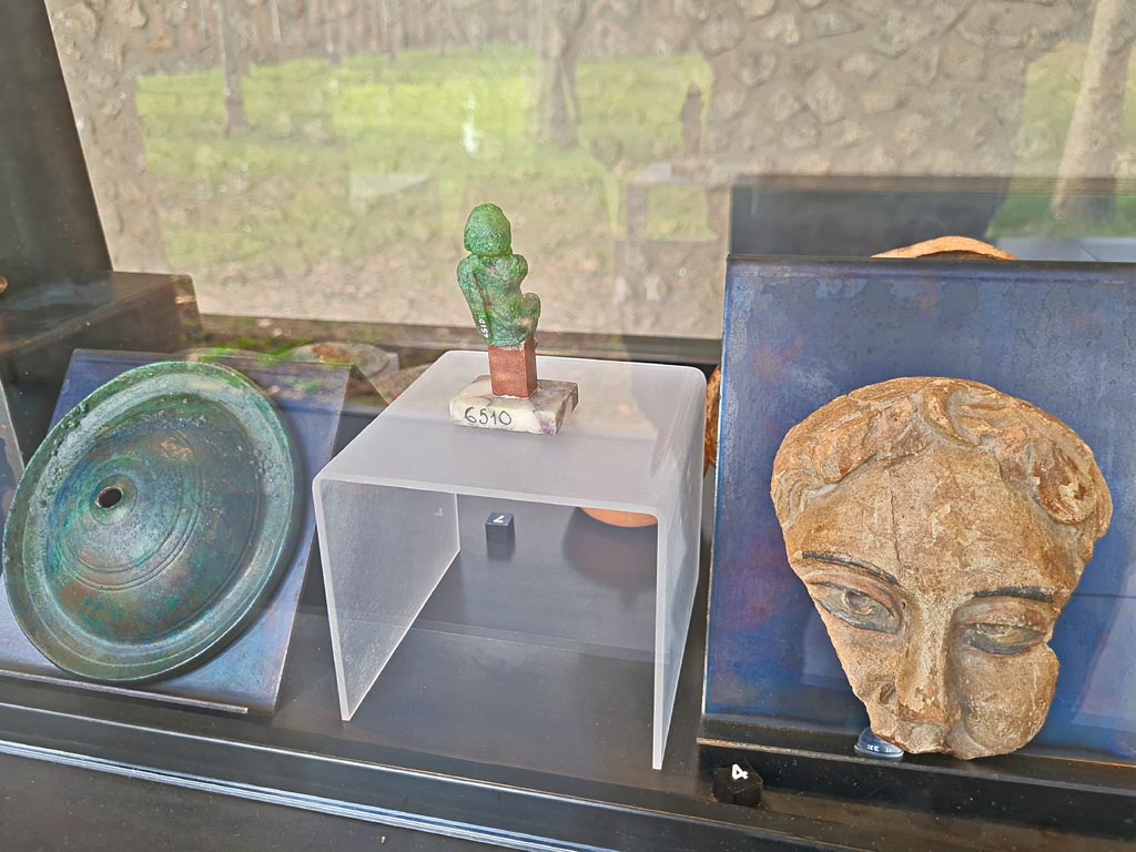 I.8.17 Pompeii. March 2024. No.7 – Rear of bronze statuette of an actor (in centre) (Ist century AD). Photo courtesy of Giuseppe Ciaramella.
Rear of cabinet, on display in exhibition in Palaestra entitled – “L’altra Pompei, vite comuni all’ombra del Vesuvio”.
On left, No.8 – Bronze cymbals from Pompeii.
On right, No.4 – one of two Terracotta theatre masks from Pompeii. 
