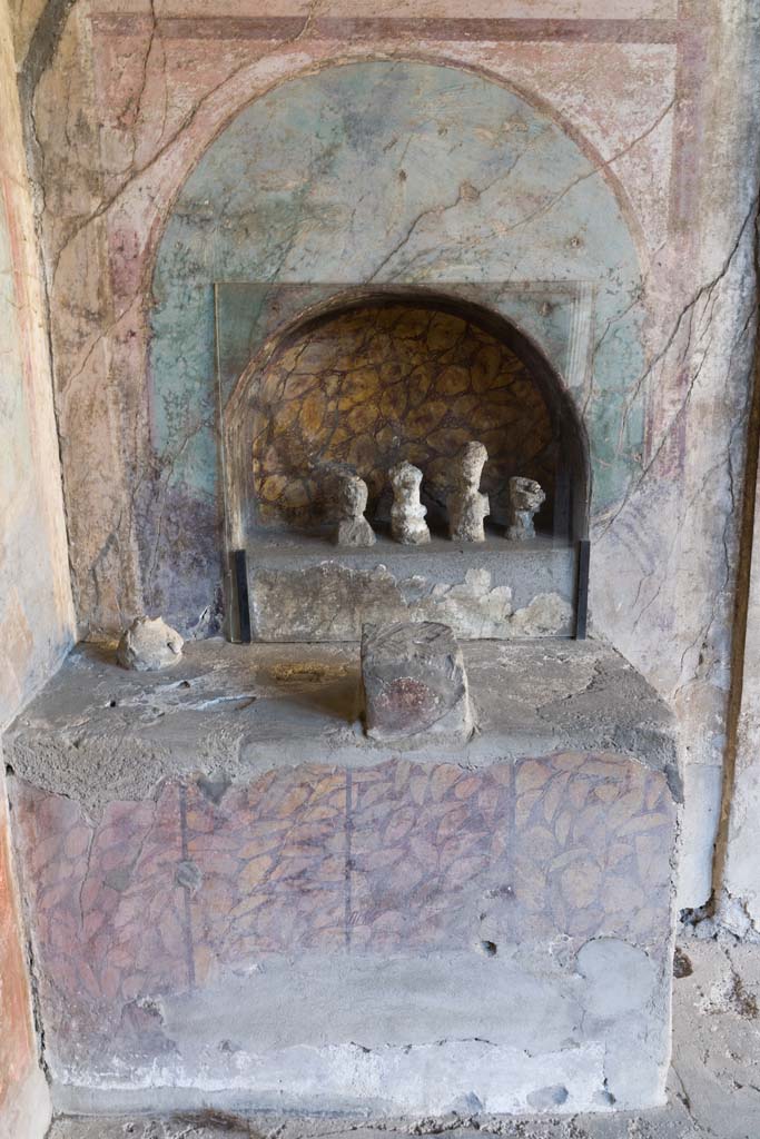 I.10.4 Pompeii. April 2022. 
Alcove 25, looking towards altar to household gods against west wall. Photo courtesy of Johannes Eber.
According to Boyce –
In the finely decorated exedra opening off the south-west corner of the peristyle is an unusual shrine.
A large rectangular, masonry podium (1.33 by 0.66, h.089) is built into the south-west corner.
In the west wall above it is a shallow arched niche (h.0.62, w.0.86, h. above the podium 0.24), set into the centre of a previously existing panel of the wall decoration. A small, rectangular, altar-like base of tufa (0.21 by 0.17, h.0.25) was built directly before the niche on the top of the podium.
The large podium is painted to represent a veneering with vertical slabs of yellow marble veined with red, placed above a dark dado.
The back wall of the niche is painted in imitation of the same marble.
The small altar-like structure is coated with red stucco.
Within the niche there stood in antiquity five figures of perishable material which left their impressions in the volcanic ash; of these impressions the excavators were able to take plaster casts. ………….
See Boyce G. K., 1937. Corpus of the Lararia of Pompeii. Rome: MAAR 14. (p.28.no. 49B).
See Giacobello, F., 2008. Larari Pompeiani: Iconografia e culto dei Lari in ambito domestico. Milano: LED Edizioni, (p.256, no.V12)

