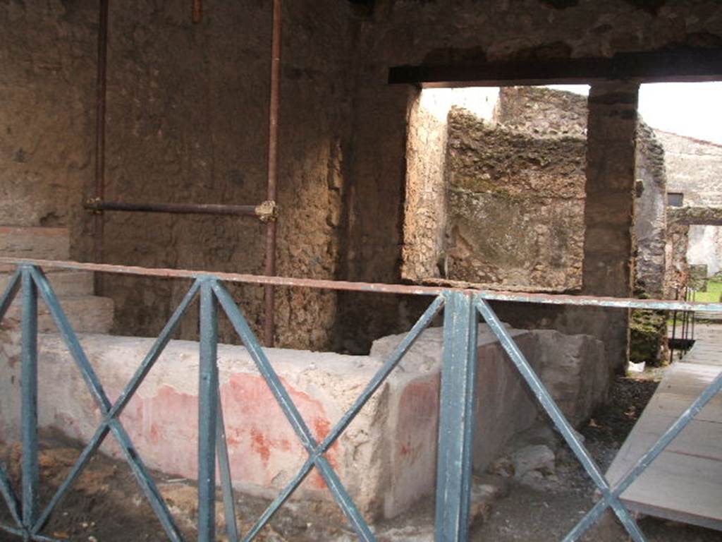 II.1.1 Pompeii. December 2004. Looking south towards two sided counter. According to NdS, the masonry of the counter was covered in stucco, which was painted to imitate a beautiful design of rich polychrome marble inlay. In the centre of the front of the counter was a yellow circle, at the sides of it were two narrow vertical rectangles. These were of white marble veined with green lozenges. At either end of the counter were two narrow vertical rectangles of yellow.  At the top, it was finished with a band of white marble.
The base was painted with a red background. The exterior side of the counter was also painted, it showed three large Iris plants with birds flying between them. At the south end of the side counter, a bronze cauldron was found. It was round with a cylindrical neck, and without a lid, it was 0.80 high. Beneath it there was a small hearth.  See Notizie di Scavi, 1917, (p.250)
