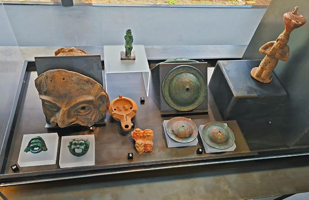 II.7.9 Pompeii. Palaestra. Some items on display in exhibition in Palaestra entitled – “L’altra Pompei, vite comuni all’ombra del Vesuvio”. 
March 2024.
No.3 – Bronze wall sconces with theatre masks, from Pompeii.
No.4 – Terracotta theatre masks (2), from Pompeii.
No.5 – Plaque with theatre masks from I.9.13.
No.6 – Oil lamp with theatre masks from I.9.9.
No.7 – Bronze statuette of an actor from I.8.17.
No.8 – Bronze cymbals from Pompeii.
No.9 – Oil lamp with Pan from I.13.2.
Photo courtesy of Giuseppe Ciaramella.
