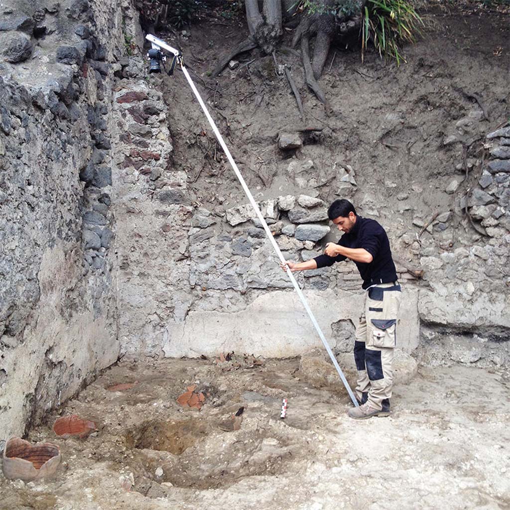 HGE28 Pompeii. 2014. Bastien Lemaire photographs the locations of potter’s wheels using a pole. Photo © Guilhem Chapelin.
According to Laetitia Cavassa, after removing the modern rubble, four circular cavities were unearthed, three of whose walls consisted of fragments of cut amphoras; these were the remains of the potter’s wheels. This was a major discovery, as it illustrated through archaeological remains what we already knew, but only with regard to frescoes. Among the rare representations from the Roman period illustrating potter’s wheels at work, two are mural paintings discovered in Pompeii. Now we have these Pompeian wheels. While these traces are fleeting, they indicate the location of the master turners as well as the how these wheels worked. We noted the presence of a central axis made of wood on which the turning wheel was placed. The entire chain of operation has now been illustrated, from the shaping of vases, unfired goblets, and the firing of vases in kilns. 
See Laetitia Cavassa, 2019. Tracing Back the Potters of Pompeii.  Article in English

HGE28 Pompeii. 2014. Bastien Lemaire photographie les emplacements des tours de potiers à l’aide d’une perche. Photo © Guilhem Chapelin.
Rapidement, après avoir enlevé les déblais modernes, nous mettons au jour quatre cavités circulaires dont la paroi est constituée, pour trois d’entre elles, par des fragments d’amphores taillées : il s’agit des vestiges des tours de potiers dont nous cherchions les emplacements. Nous les avons enfin trouvés.
La découverte est de taille : elle vient illustrer par des traces archéologiques ce que nous ne connaissions jusqu’alors que par des fresques. Parmi les rares représentations d’époque romaine illustrant des potiers en train de travailler, deux sont des peintures murales découvertes à Pompéi. Maintenant, nous avons les tours pompéiens. Ces traces, bien que fugaces, indiquent la position des maîtres-tourneurs ainsi que le fonctionnement de ces tours de potiers. On constate la présence d’un axe central en bois sur lequel était placée la roue du tour. Toute la chaîne opératoire est désormais illustrée, de la mise en forme des vases, en passant par les gobelets crus, jusqu’à la cuisson des vases dans les fours.
Voir Laetitia Cavassa, 2019. Sur la trace des potiers de Pompéi.   Article en français 
